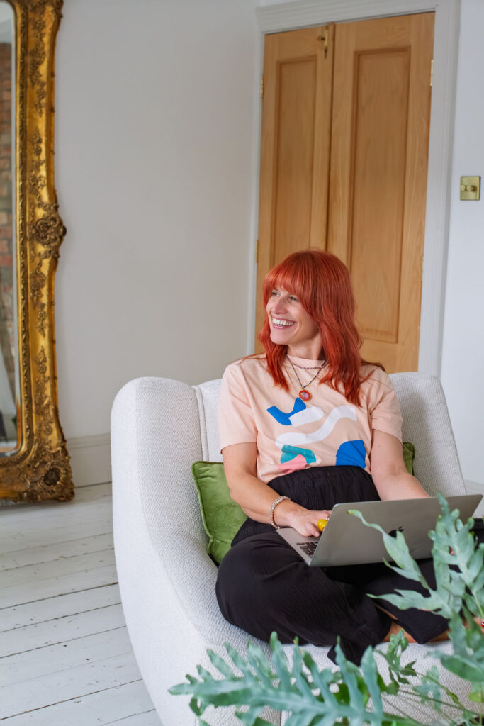 Nicola smiling on a white armchair with her laptop, beside a gold mirror