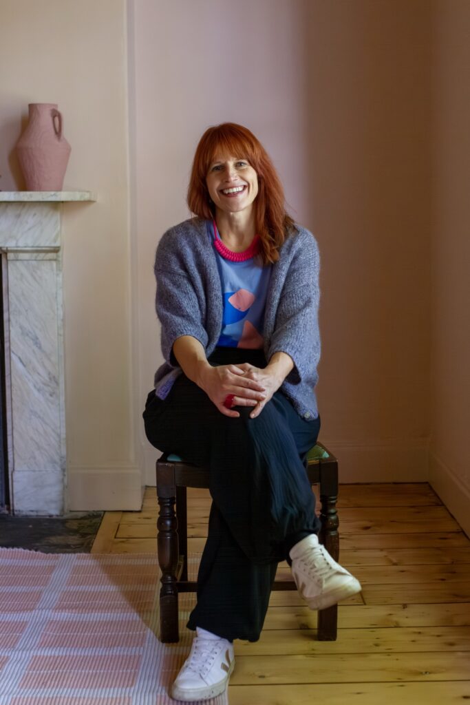 Nicola smiling, sitting on a wooden chair in a pink room